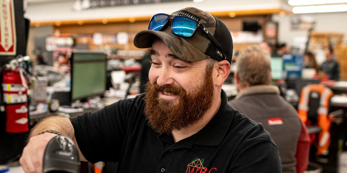 A bearded male employee in a black polo shirt with WBC logo is working at the checkout counter, using a handheld barcode scan