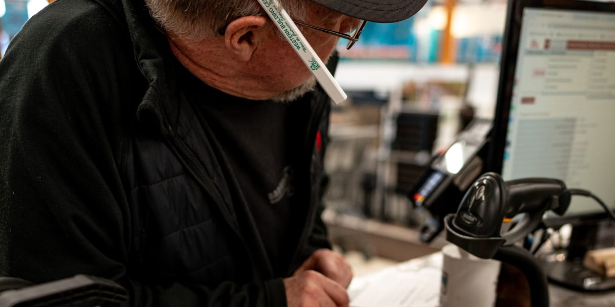 A Western Building Center employee wearing a black hoodie, baseball cap, and glasses is working at a service counter, examini