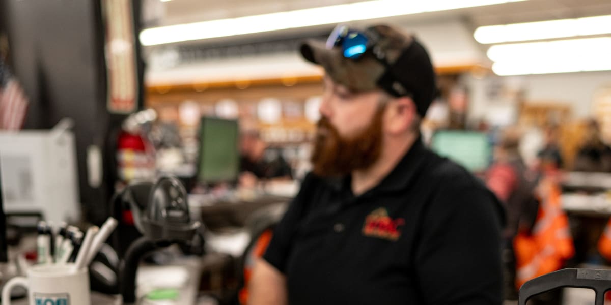A bearded employee wearing a cap and dark polo shirt with company logo sits at what appears to be a service counter or orderi