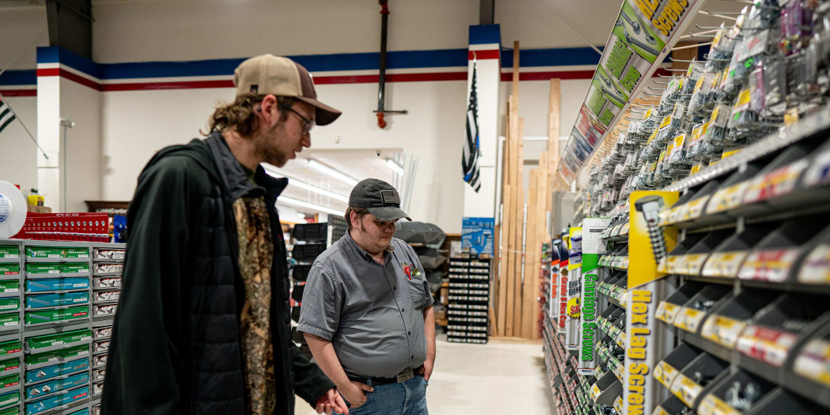 Two men browse tools and hardware in a store aisle, with one customer in dark clothing and baseball cap looking at products w