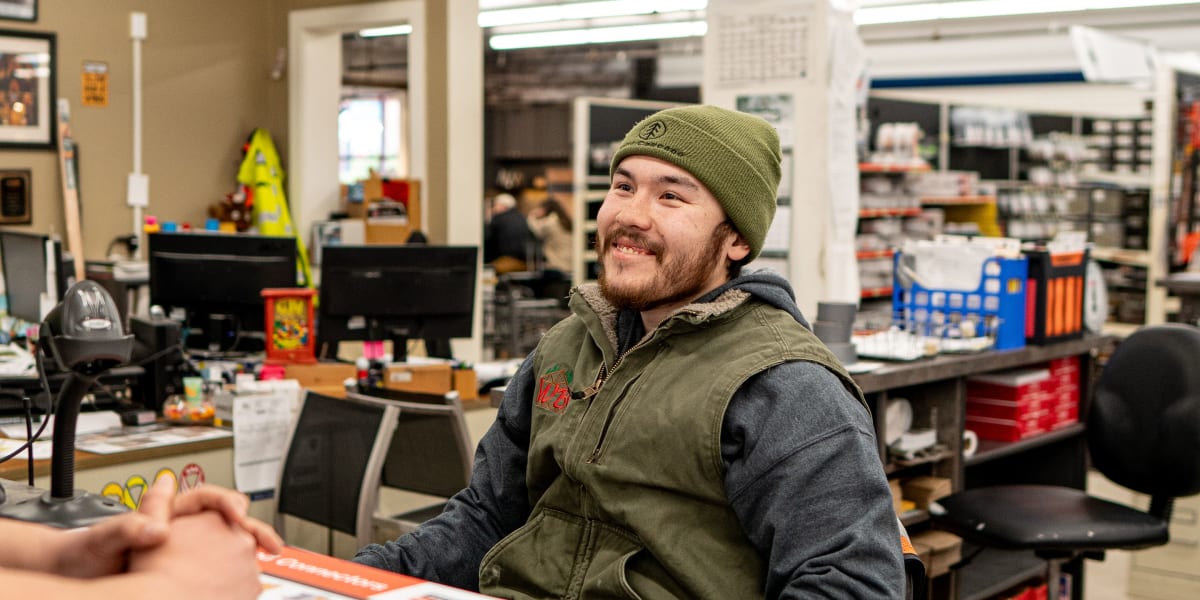 A smiling male employee wearing a green knit beanie and olive vest is sitting at the contractor's service center counter, eng