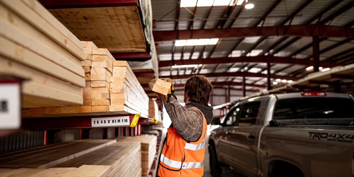 A worker in an orange high-visibility safety vest is loading dimensional lumber from warehouse shelving into a Toyota TRD pic