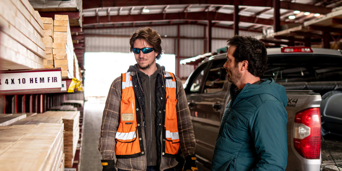 Two men having a conversation in a lumber warehouse, with one wearing a high-visibility orange safety vest, work boots, and s