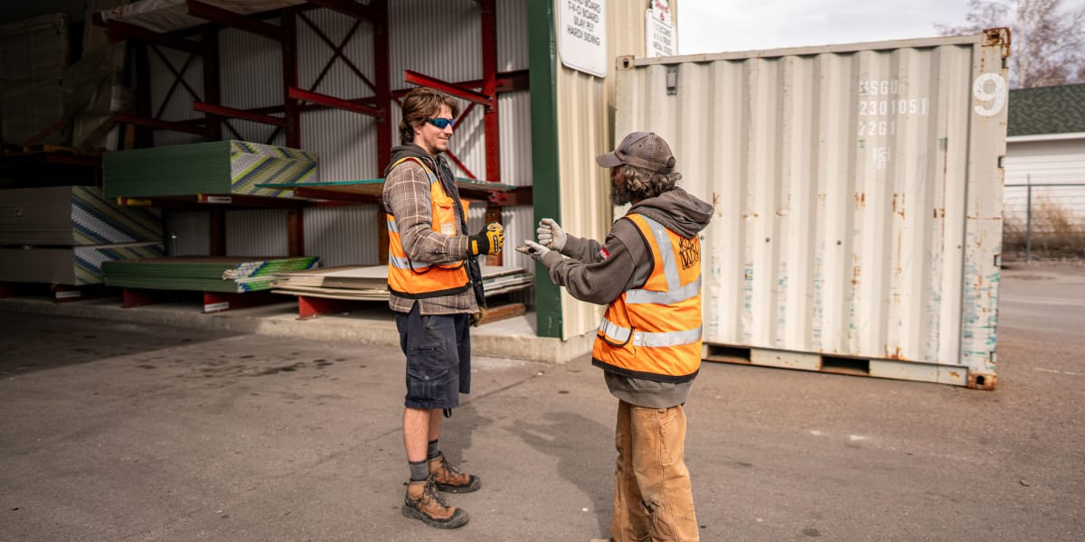 Two construction workers in high-visibility orange safety vests are having a conversation in an industrial lumber yard settin