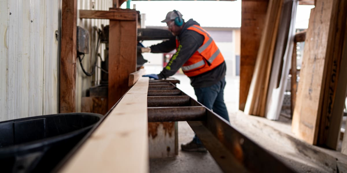 A construction worker in an orange high-visibility safety vest and hard hat is operating what appears to be a large planer or