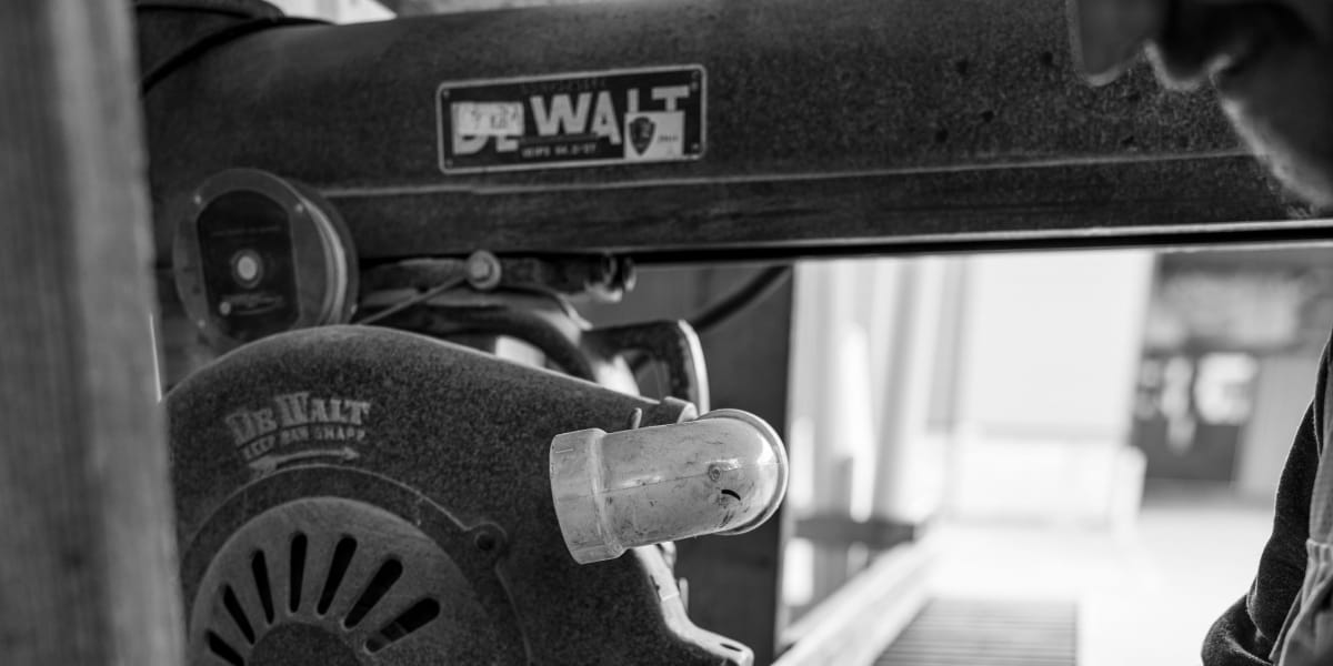 Black and white image showing a person using a DeWalt radial arm saw to cut wooden boards in a lumber yard workshop setting