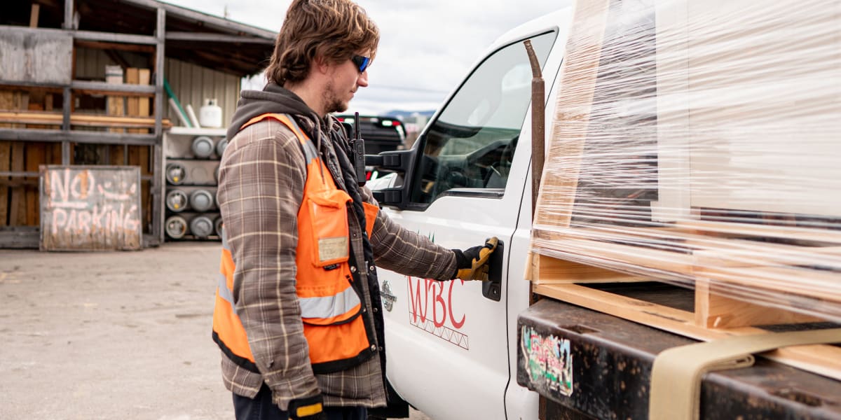 A construction worker in an orange safety vest and work boots is opening the door of a white work truck in what appears to be