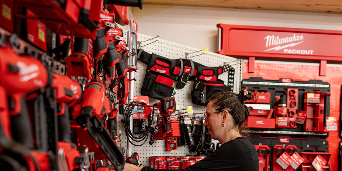 A female employee in a black shirt is helping organize or restock Milwaukee power tools on a pegboard display wall in the too