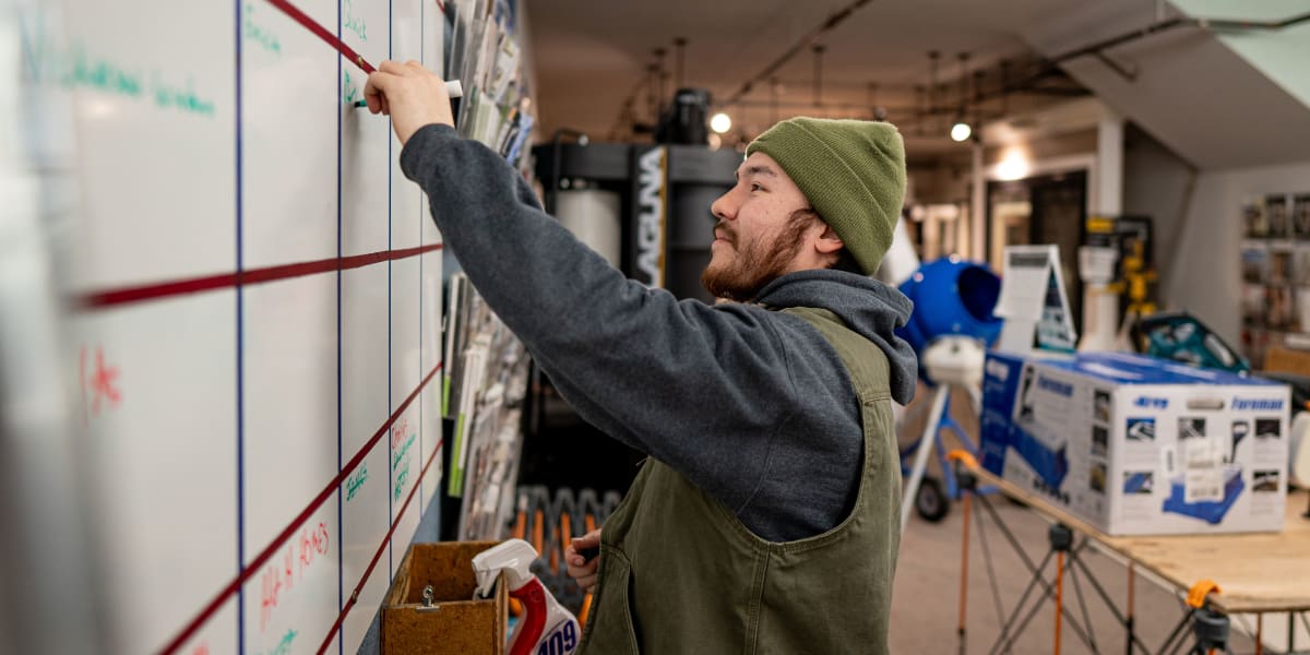 An employee wearing a green knit beanie and gray hoodie is pointing at or drawing on a large whiteboard covered with colored