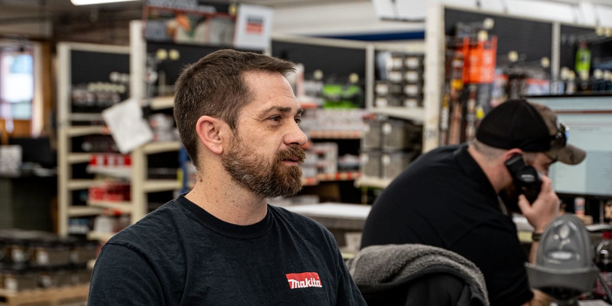 Two employees working in a building supply store interior, with one bearded man in a black Makita t-shirt sitting at what app
