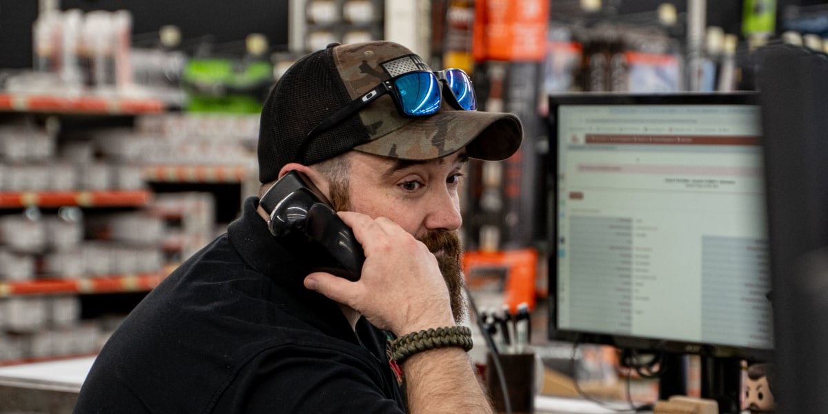 An employee at Western Building Center wearing a camouflage baseball cap with sunglasses sits at a desk talking on a corded p