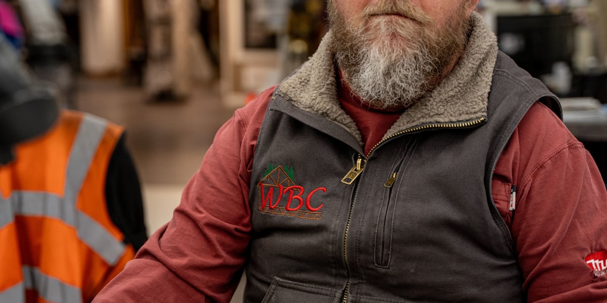 A bearded employee wearing a WBC (Western Building Center) branded brown vest over a red long-sleeve shirt sits at what appea