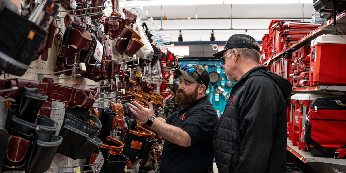 Two men are shopping in the tool belt and leather goods aisle of a hardware store, examining various tool pouches, holsters,