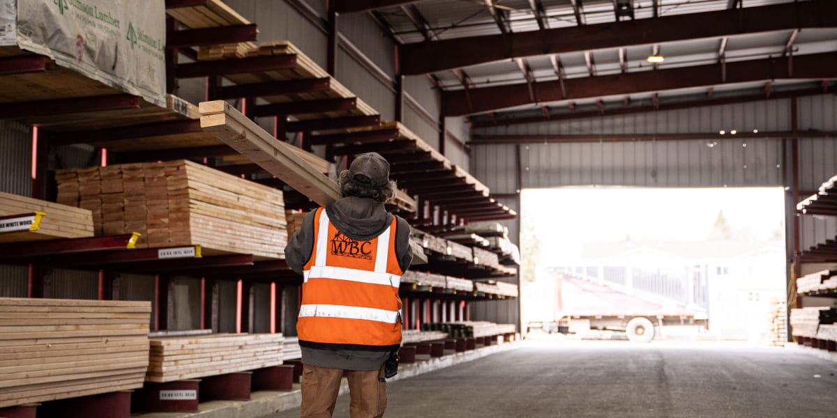 A worker wearing an orange high-visibility safety vest with reflective stripes walks through a large lumber warehouse