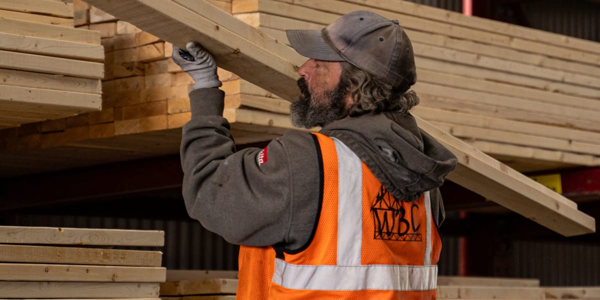 A construction worker in a high-visibility orange safety vest and baseball cap is handling dimensional lumber in a covered lu