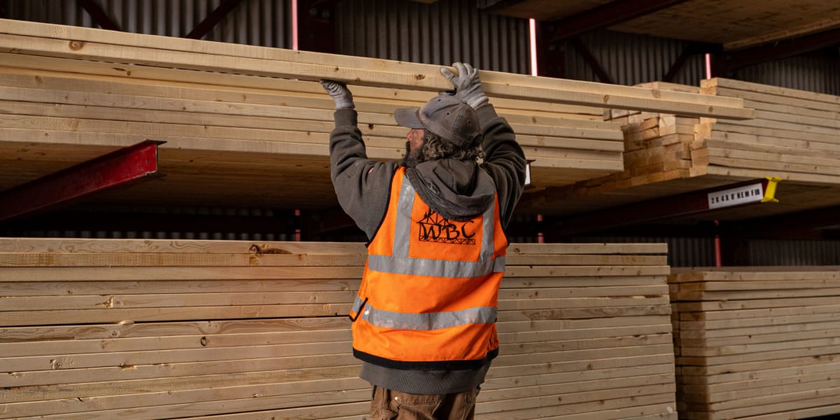 A worker in an orange safety vest and WPC company logo is selecting lumber from organized stacks in a covered lumber yard