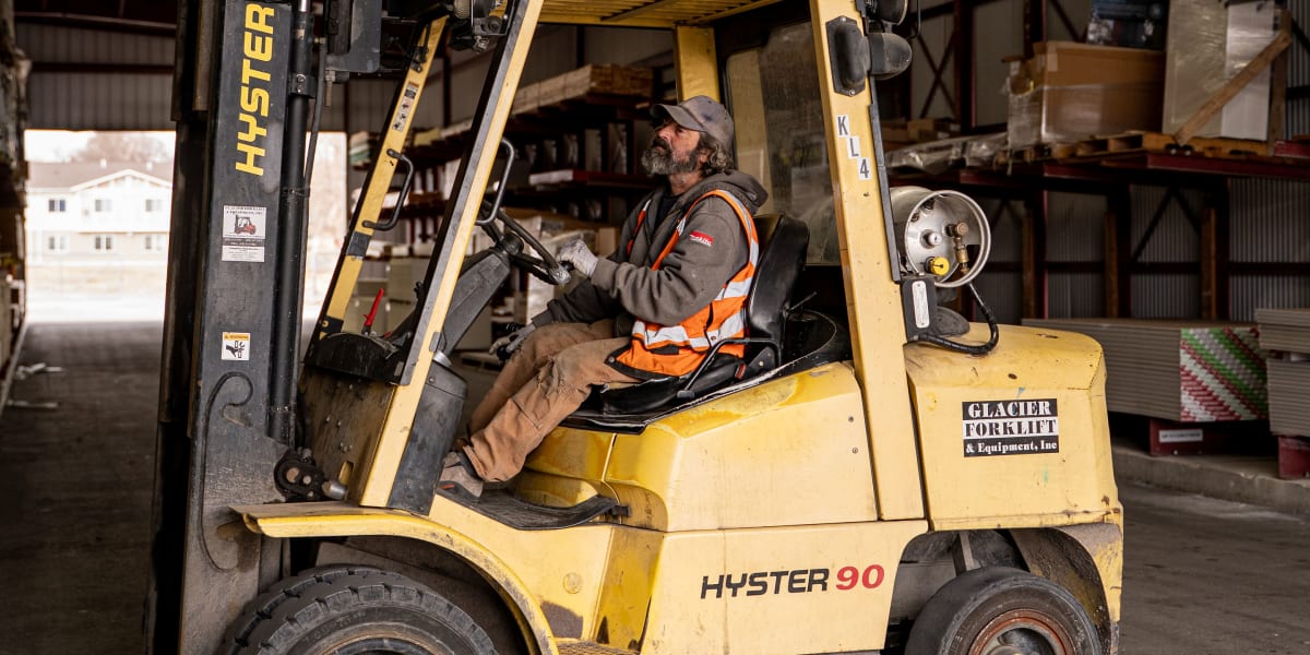A forklift operator wearing a high-visibility orange safety vest and baseball cap sits in a yellow Hyster 90 forklift inside