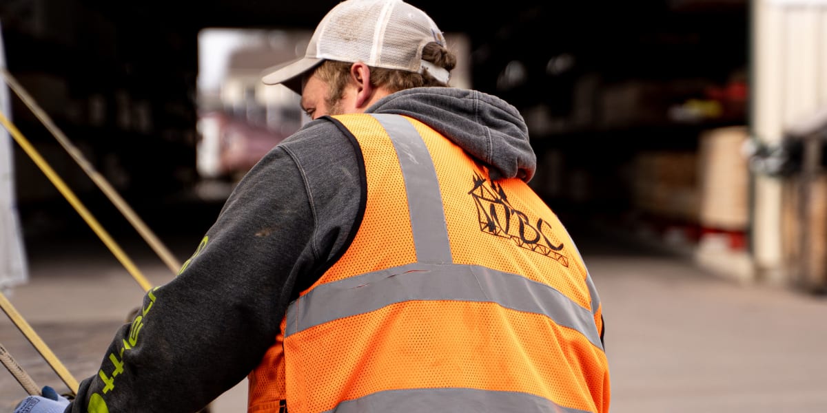 A worker in a high-visibility orange safety vest with reflective stripes is working at what appears to be a lumber yard or bu