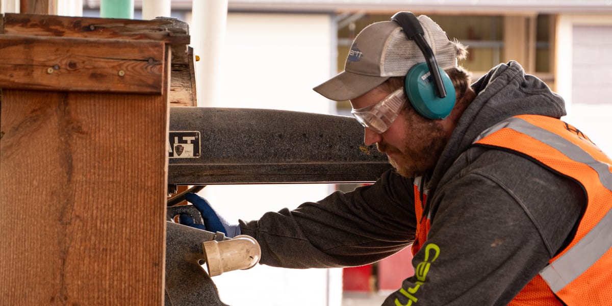 A construction worker wearing a high-visibility orange safety vest and protective ear muffs operates a large power saw or cut