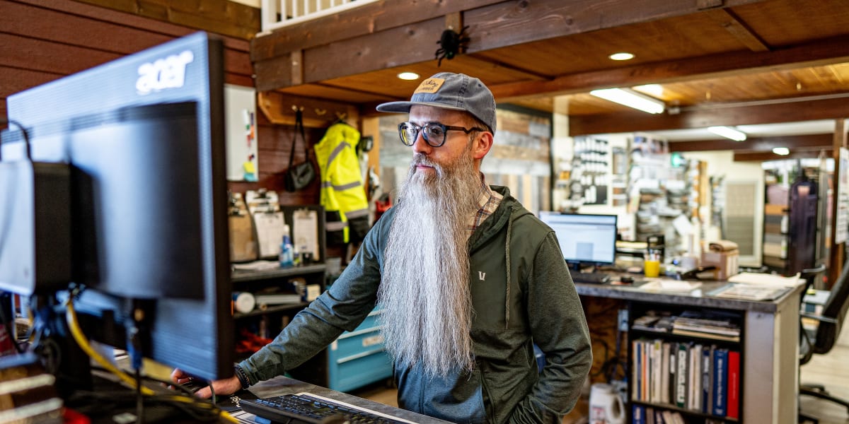 A bearded employee wearing glasses, a baseball cap, and olive green shirt sits at a computer workstation in what appears to b