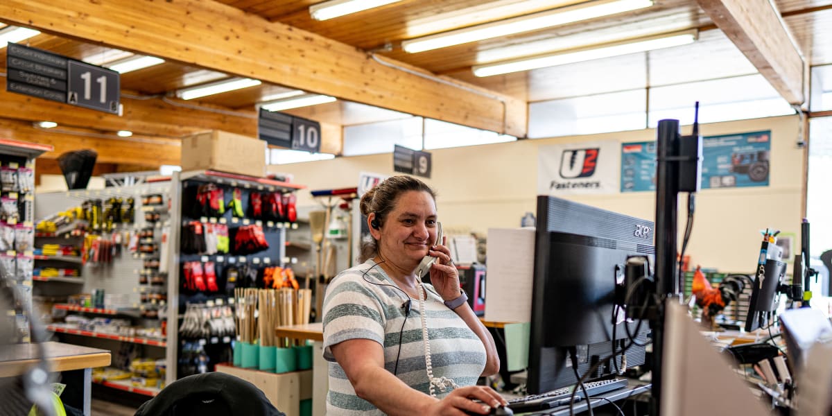 A female employee wearing a striped shirt is working at the store checkout counter, talking on the phone while looking at a c