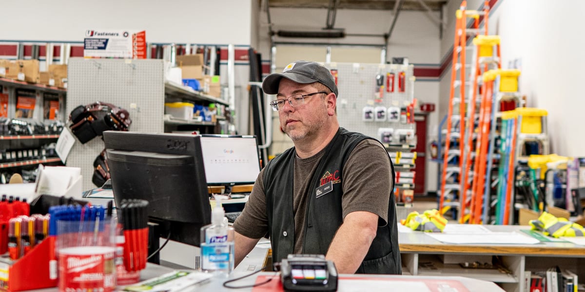 An employee at the customer service counter in the store, wearing glasses, a cap, and a dark polo shirt with company logo