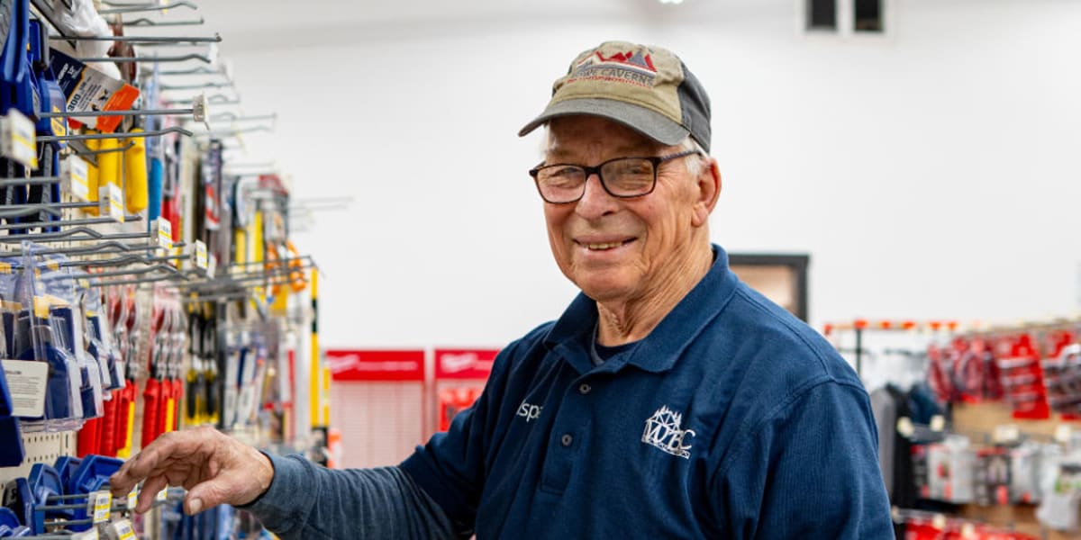 A smiling elderly male employee wearing glasses, a camouflage baseball cap, and a navy blue Western Building Center polo shir