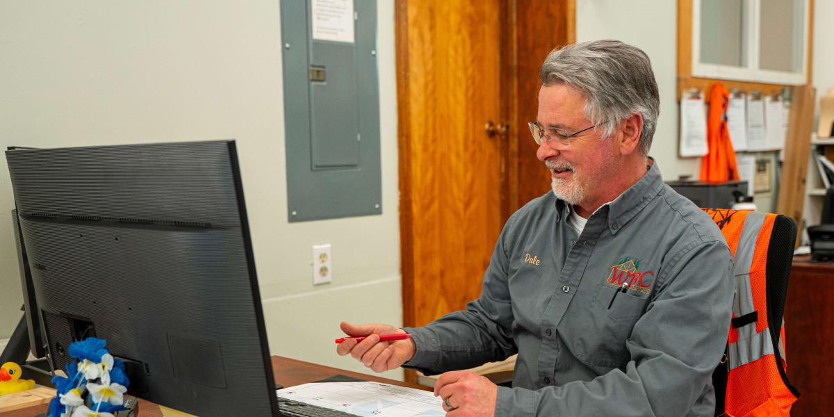 A middle-aged man with gray hair and beard sits at his office desk, working at a computer monitor while reviewing paperwork