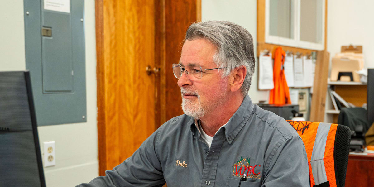 A middle-aged man named Dale wearing glasses and a gray work shirt with WBC (Western Building Center) logo sits at a wooden d