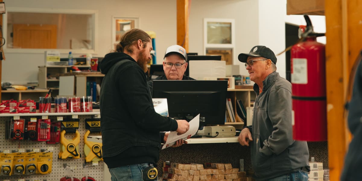 Three men are engaged in conversation at the checkout counter of a building supply store, with one bearded customer in a blac