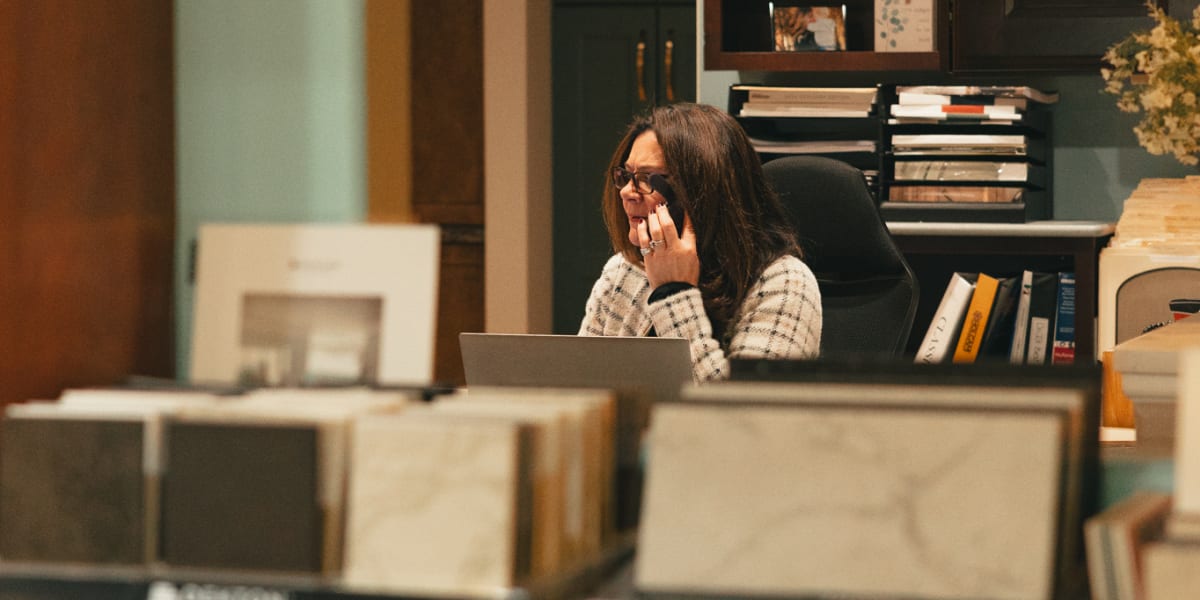 A woman in a checkered shirt sits at a desk in what appears to be an office setting, speaking on the phone while working at a