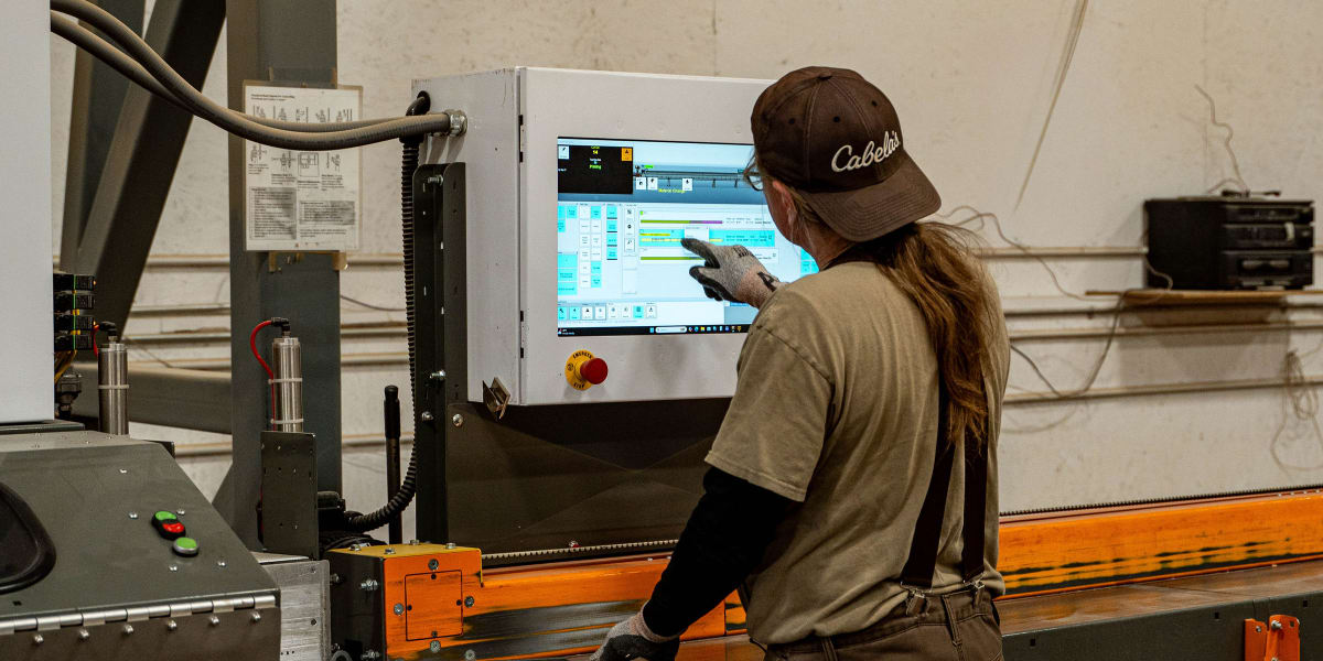A female employee wearing a brown Cabela's cap and work clothes operates a computerized truss manufacturing machine in an ind