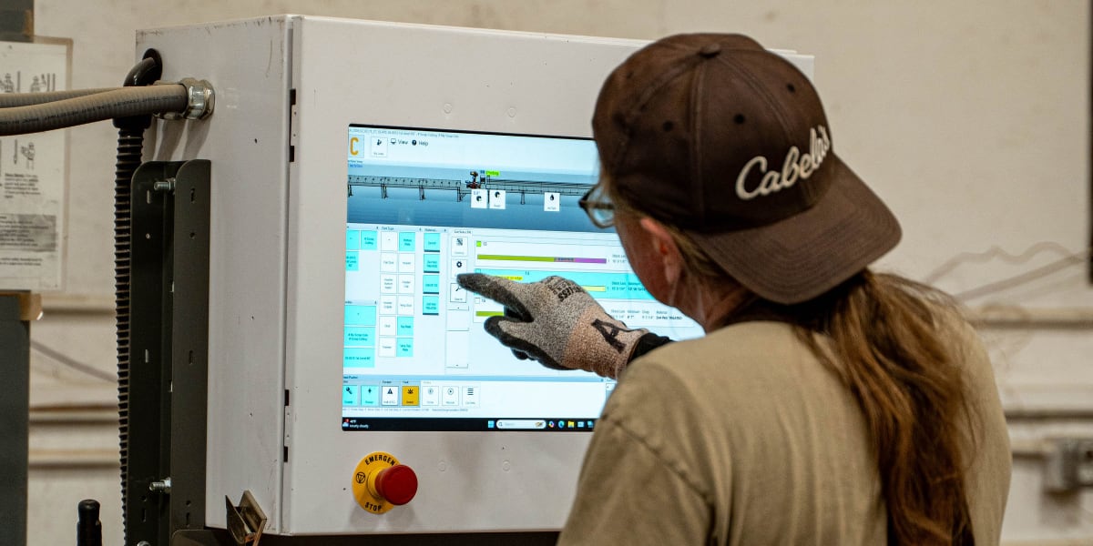 A worker in work attire and a Cabela's baseball cap operates computerized truss manufacturing equipment in an industrial sett