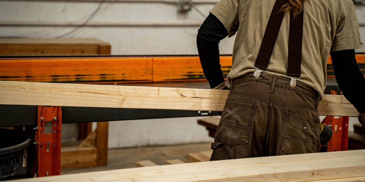 A woodworker in brown overalls and suspenders is working with dimensional lumber at an orange workbench in what appears to be