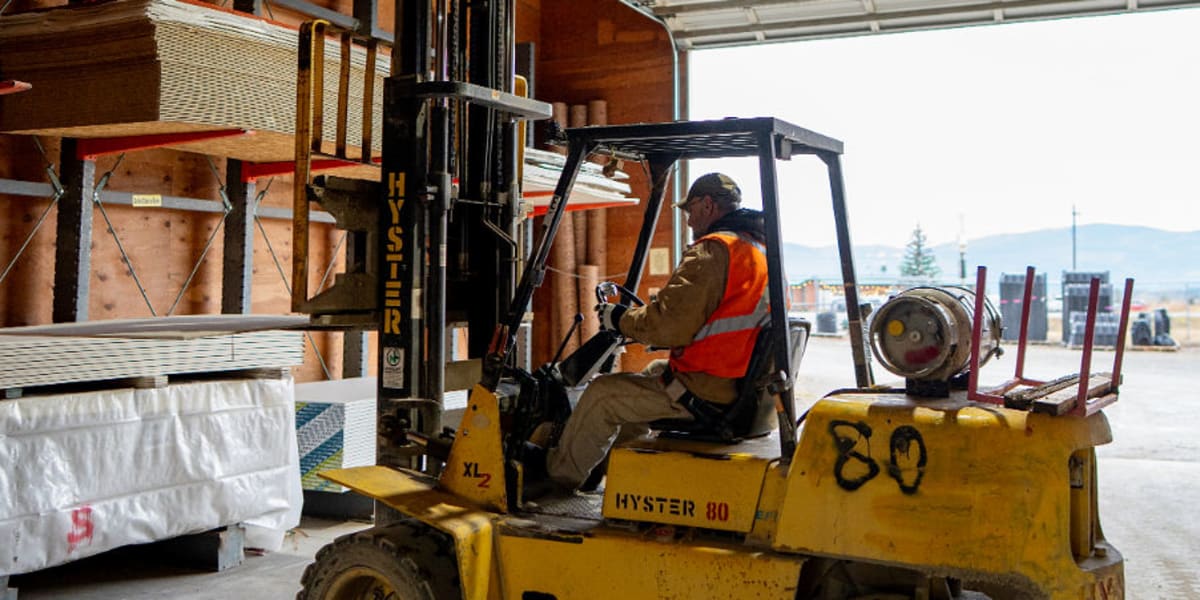 A yellow Hyster 80 forklift is operating inside a warehouse facility, positioned next to palletized building materials and lu