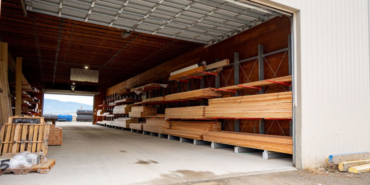 Interior view of a covered lumber storage warehouse showing organized racks of dimensional lumber and building materials