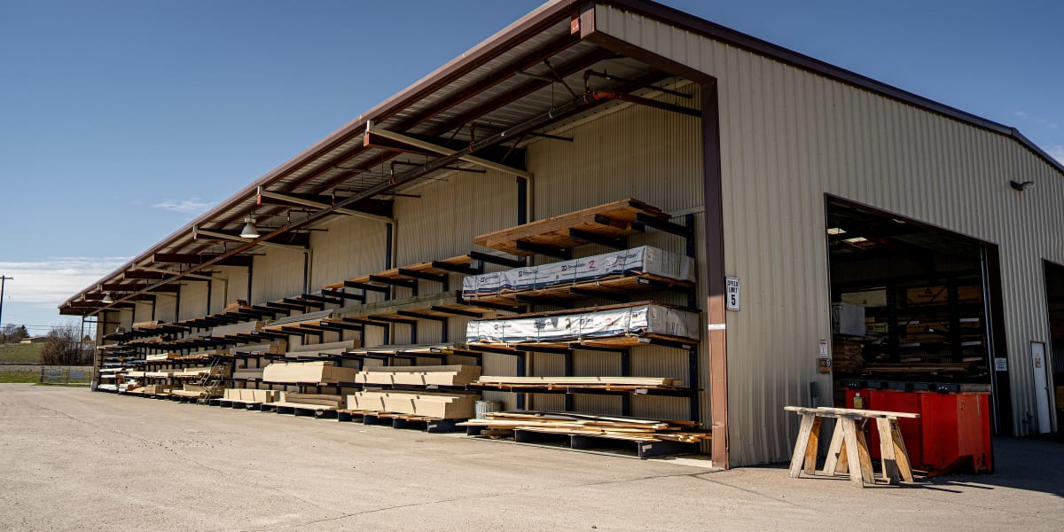 This image shows the exterior of a large industrial building supply warehouse with extensive lumber storage racks extending a