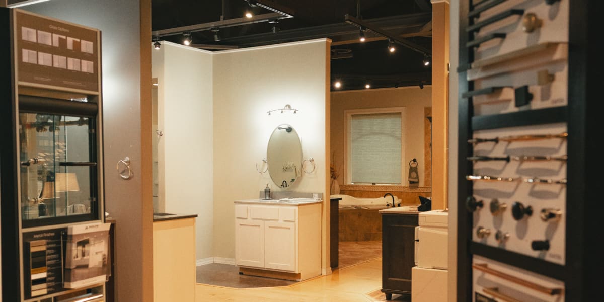 Interior showroom view of a building supply store featuring a kitchen and bath display area with white cabinetry, exposed cei