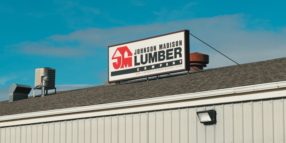 Exterior view of a Johnson Madison Lumber Company building showing the white metal siding facade and prominent rooftop signag