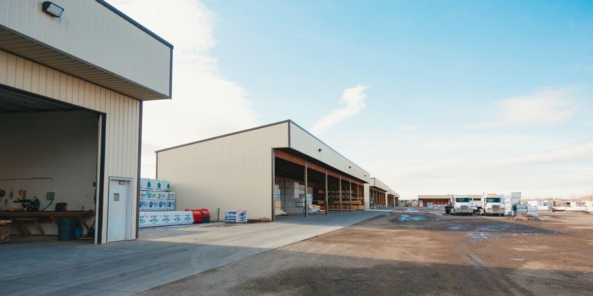 Wide exterior view of an industrial building supply facility showing large metal warehouse buildings with concrete loading ar