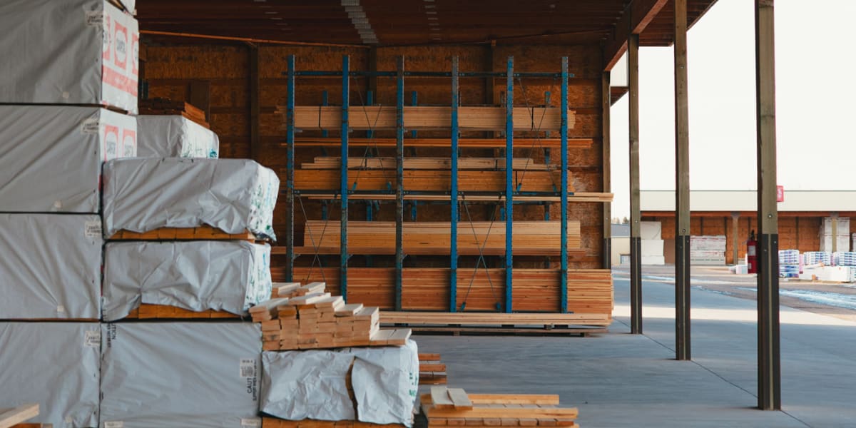 Interior view of a lumber warehouse showing stacked lumber packages wrapped in white protective covering on the left side and