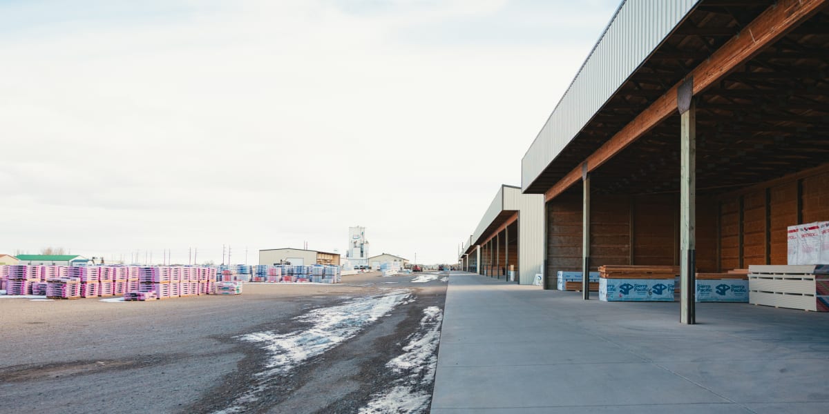 A wide view of a modern lumber yard warehouse facility showing a large industrial building with metal siding and exposed wood