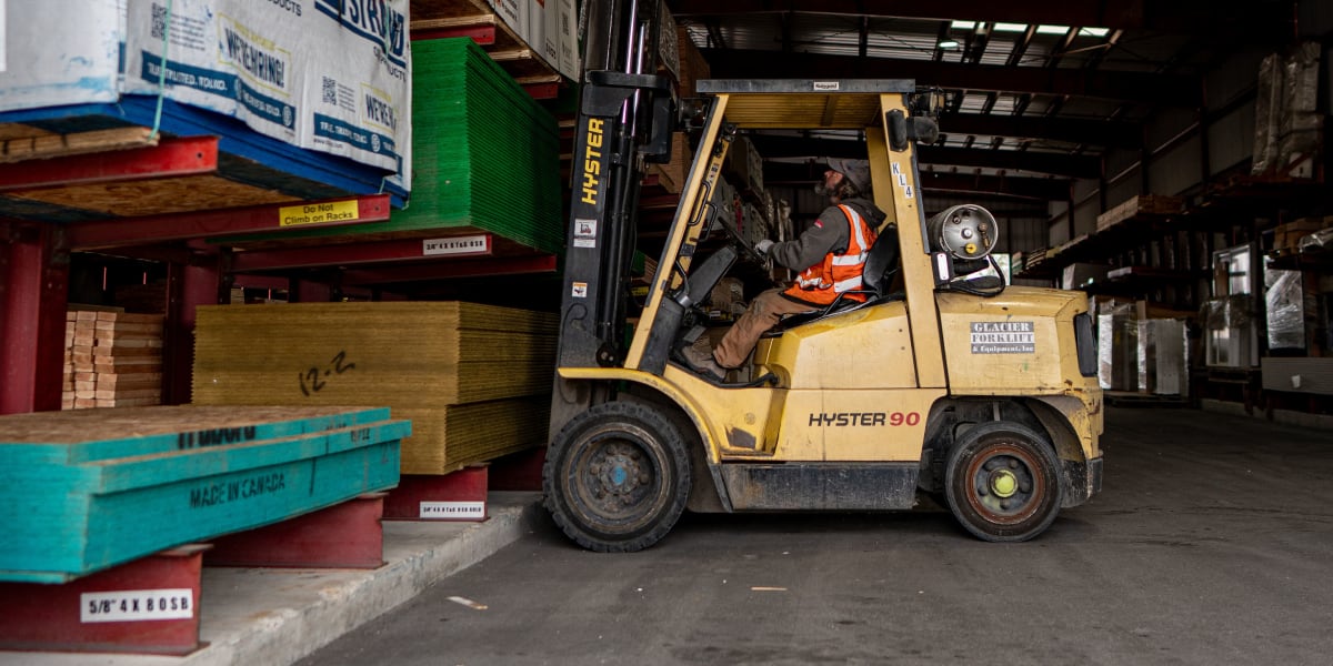 A forklift operator in safety vest is loading or organizing building materials in the warehouse section of the lumber yard