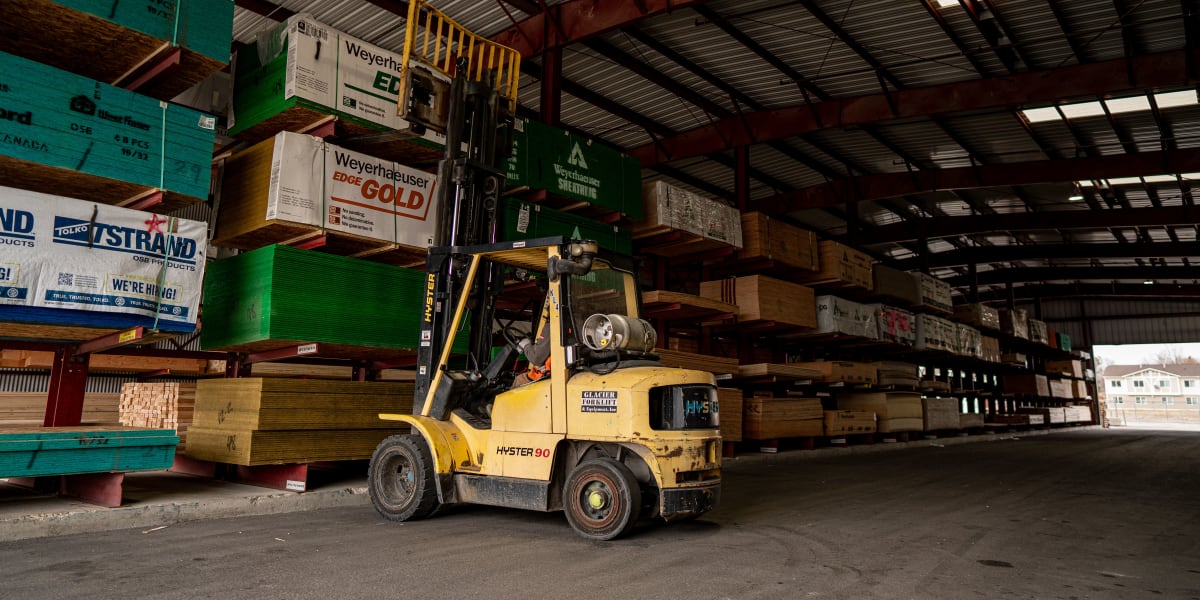 Interior view of a large warehouse lumber yard showing a yellow Hyster 90 forklift positioned among towering racks of lumber