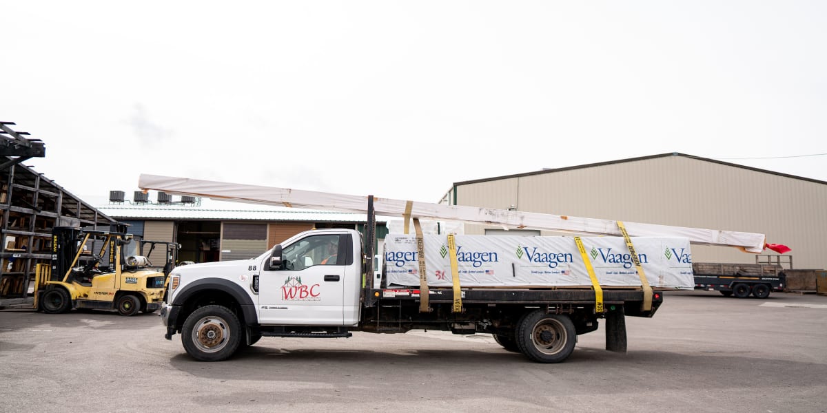 A white WBC delivery truck with flatbed trailer loaded with Vaagen lumber products is parked at an industrial lumber yard fac