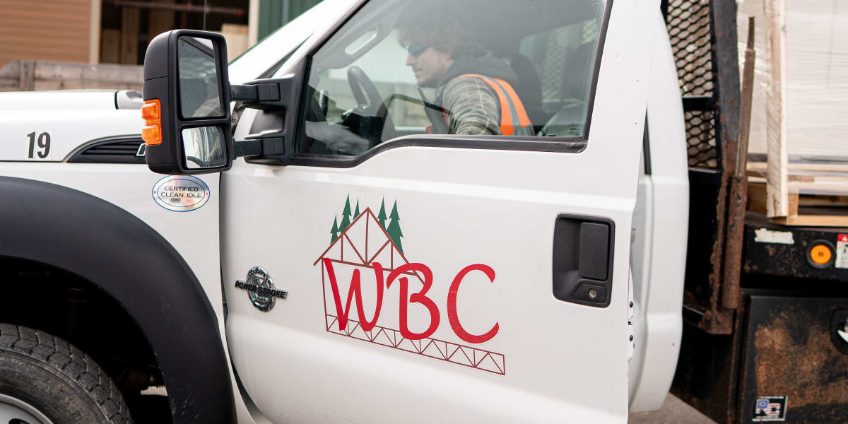 A white WBC delivery truck with the company logo and green tree graphics is parked at a lumber yard or construction site