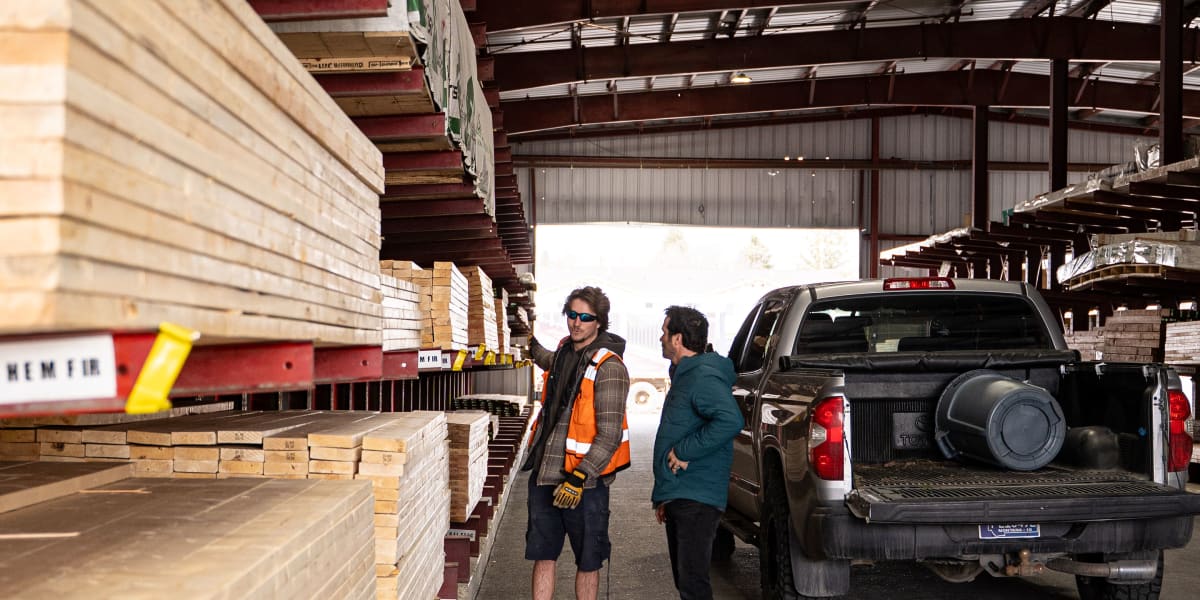 Two people are standing in a covered lumber yard warehouse area next to a dark pickup truck