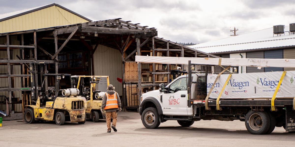 A busy lumber yard scene at Western Building Center showing industrial operations with a yellow forklift, delivery truck with