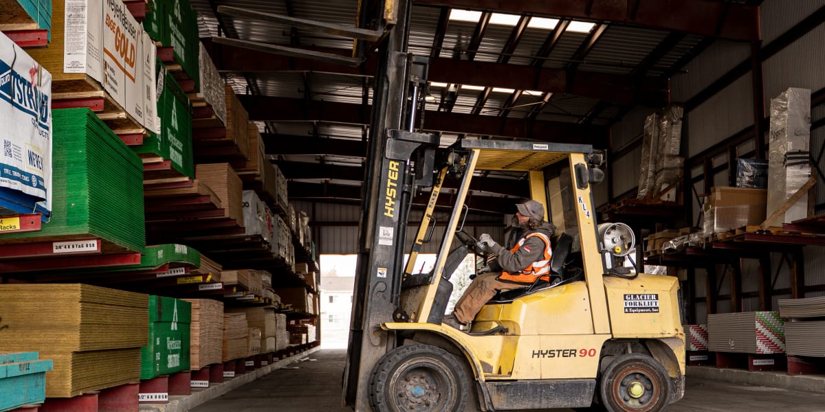 Interior warehouse view showing a yellow Hyster 90 forklift operated by a worker in an orange safety vest navigating between
