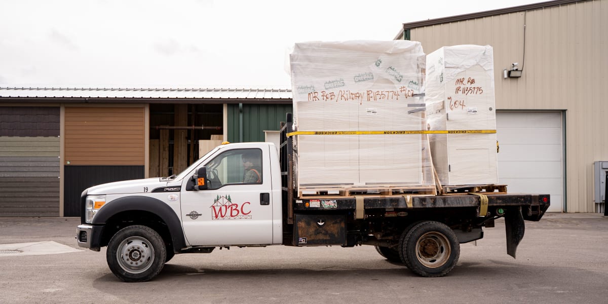 A white WBC delivery truck with flatbed is parked outside a warehouse facility, loaded with wrapped building materials on pal